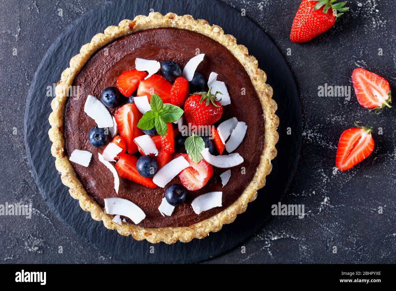Crostata francese con ripieno di cioccolato fondente, decorata con fragole e mirtilli, patatine di cocco sulla parte superiore, servite su fondo di cemento scuro Foto Stock
