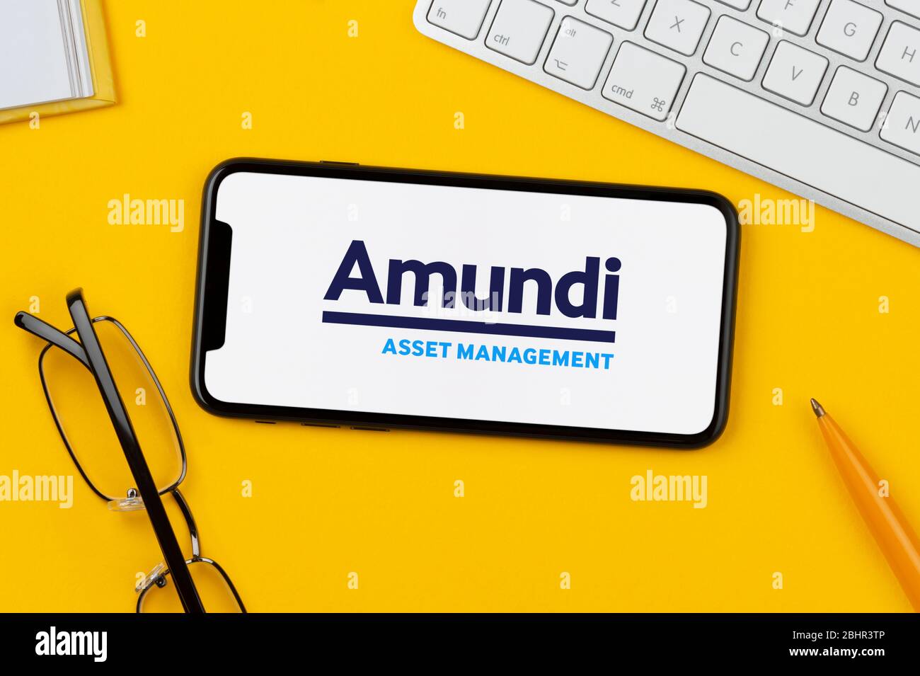 Uno smartphone con il logo Amundi Asset Management è posizionato su uno sfondo giallo insieme a tastiera, occhiali, penna e libro (solo per uso editoriale). Foto Stock