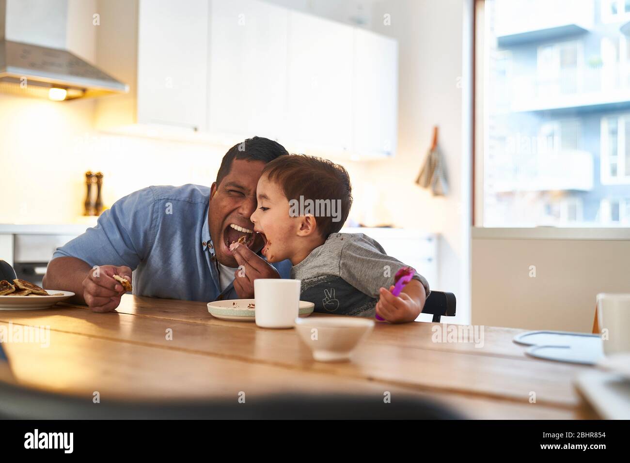 Un uomo e un figlio a un tavolo da cucina entrambi con bocche spalancate a mangiare un pezzo di torta al cioccolato. Foto Stock