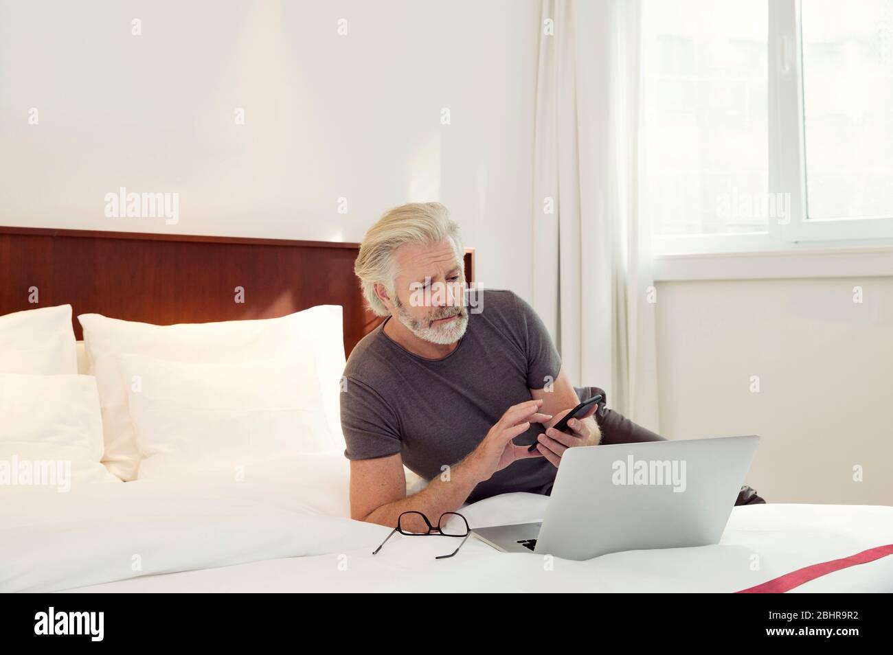 Un uomo sdraiato su un letto che guarda il suo laptop e il suo telefono cellulare. Foto Stock