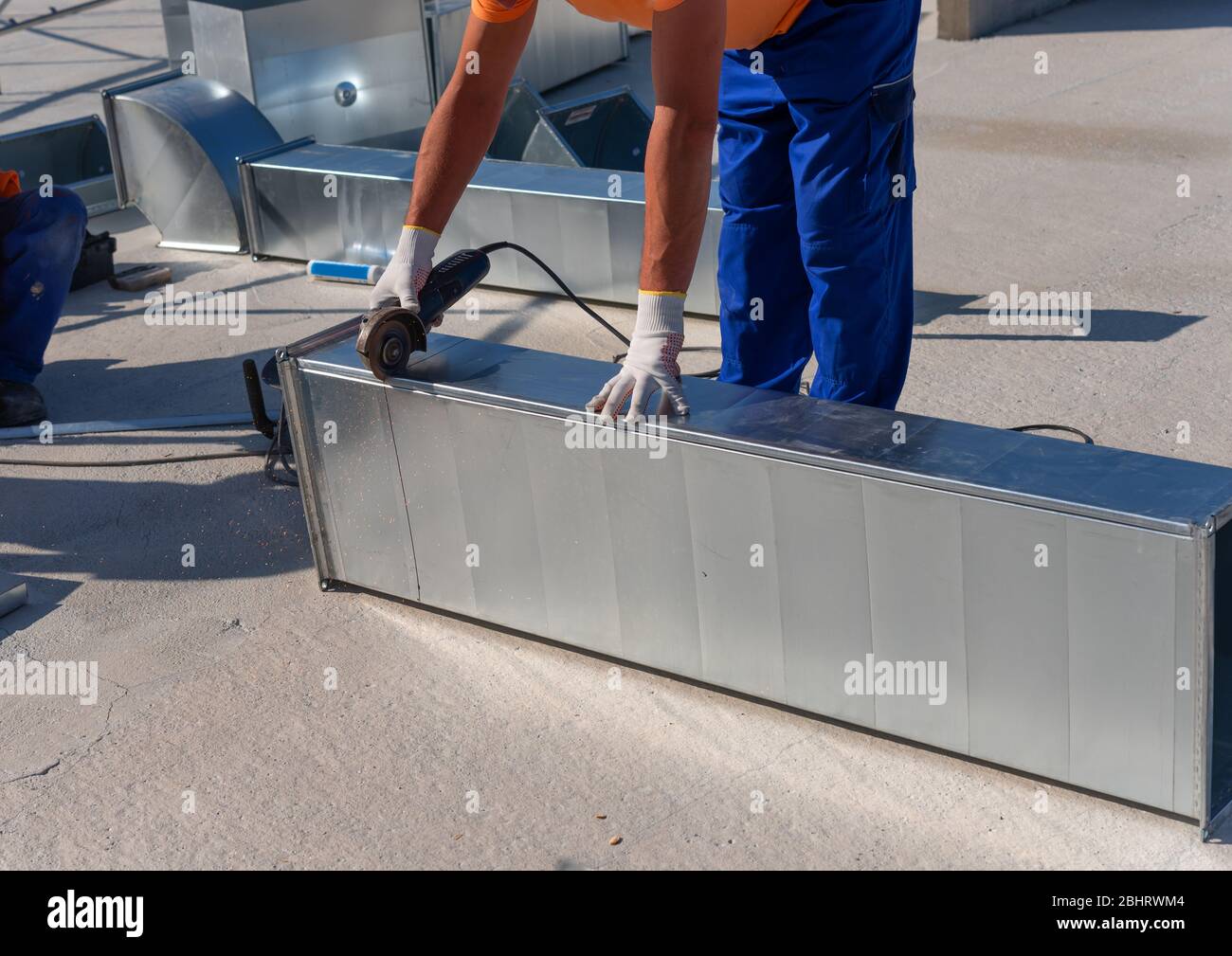 Il tecnico HVAC sta lavorando su un tetto di nuovo edificio industriale. Vista ravvicinata del giovane tecnico che ripara un condotto d'aria con la smerigliatrice angolare Foto Stock