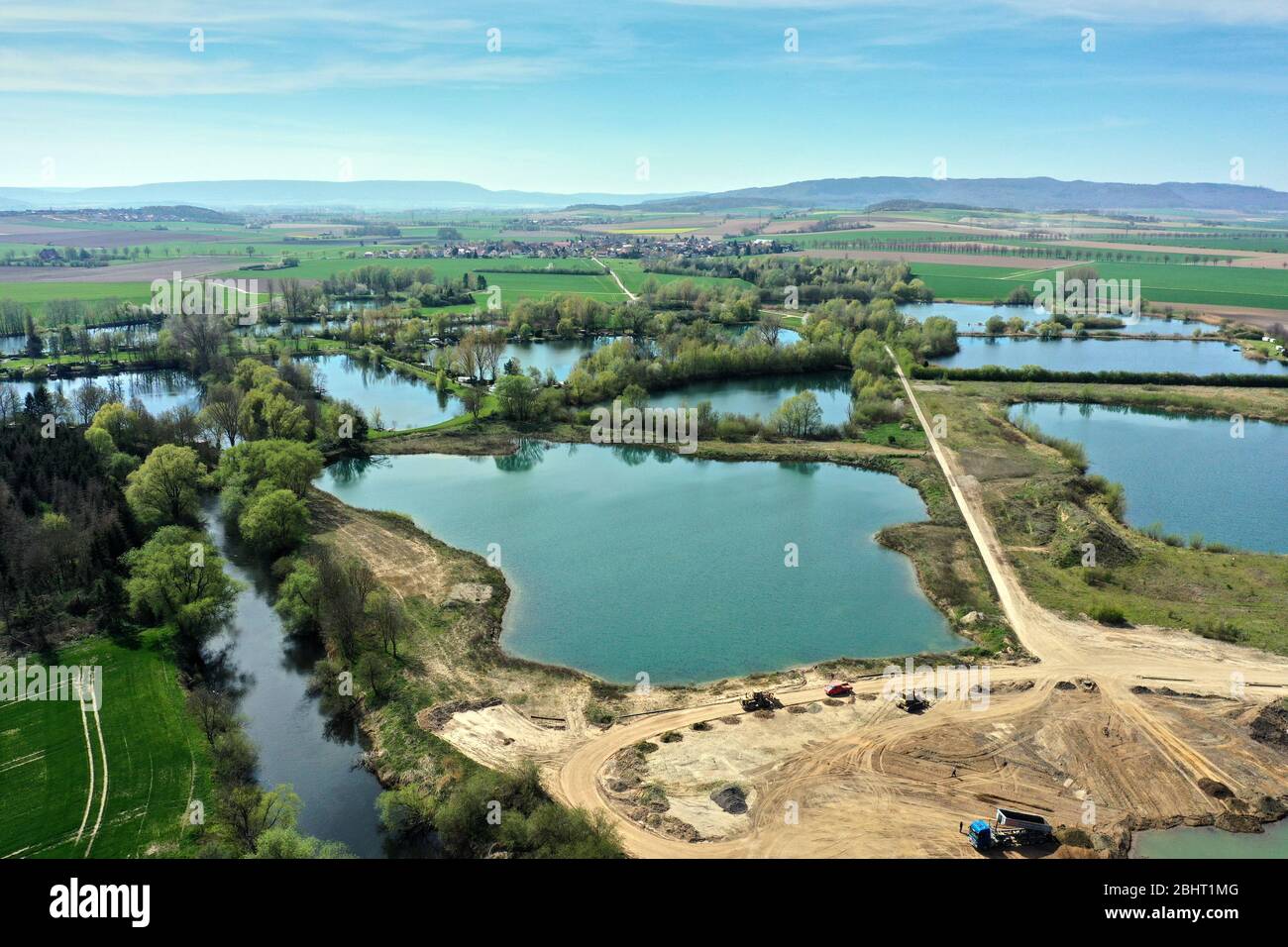 Veduta aerea di un attivo stagno ghiaia accanto al fiume Leine vicino Sarstedt, Germania, con un camion e mucchi di sabbia e un villaggio sullo sfondo Foto Stock