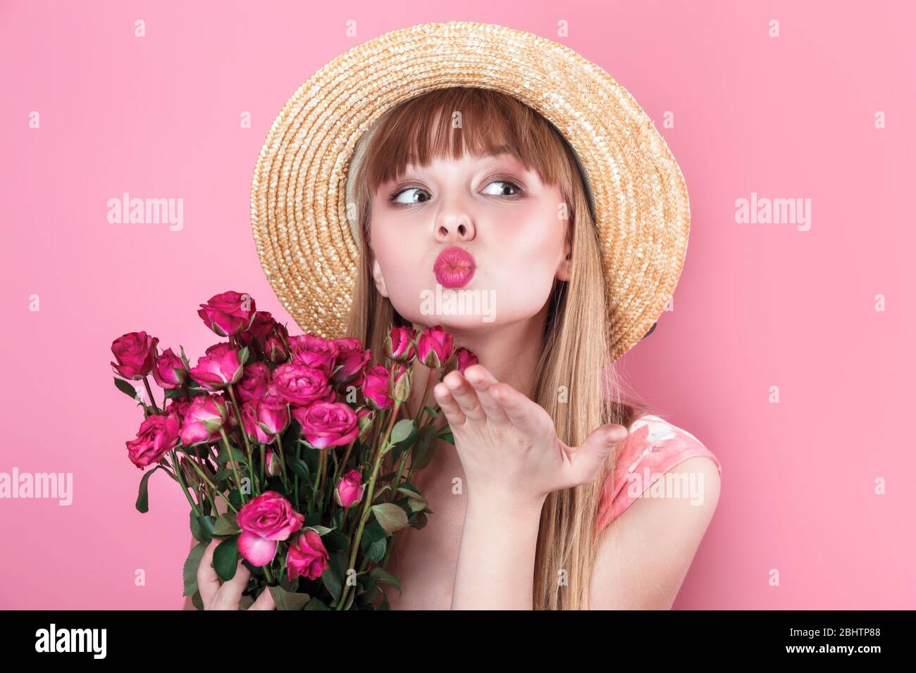 Giovane donna sorridente in estate vestito e cappello di paglia tenendo bouquet di rose e soffiando un bacio con mano su aria Foto Stock
