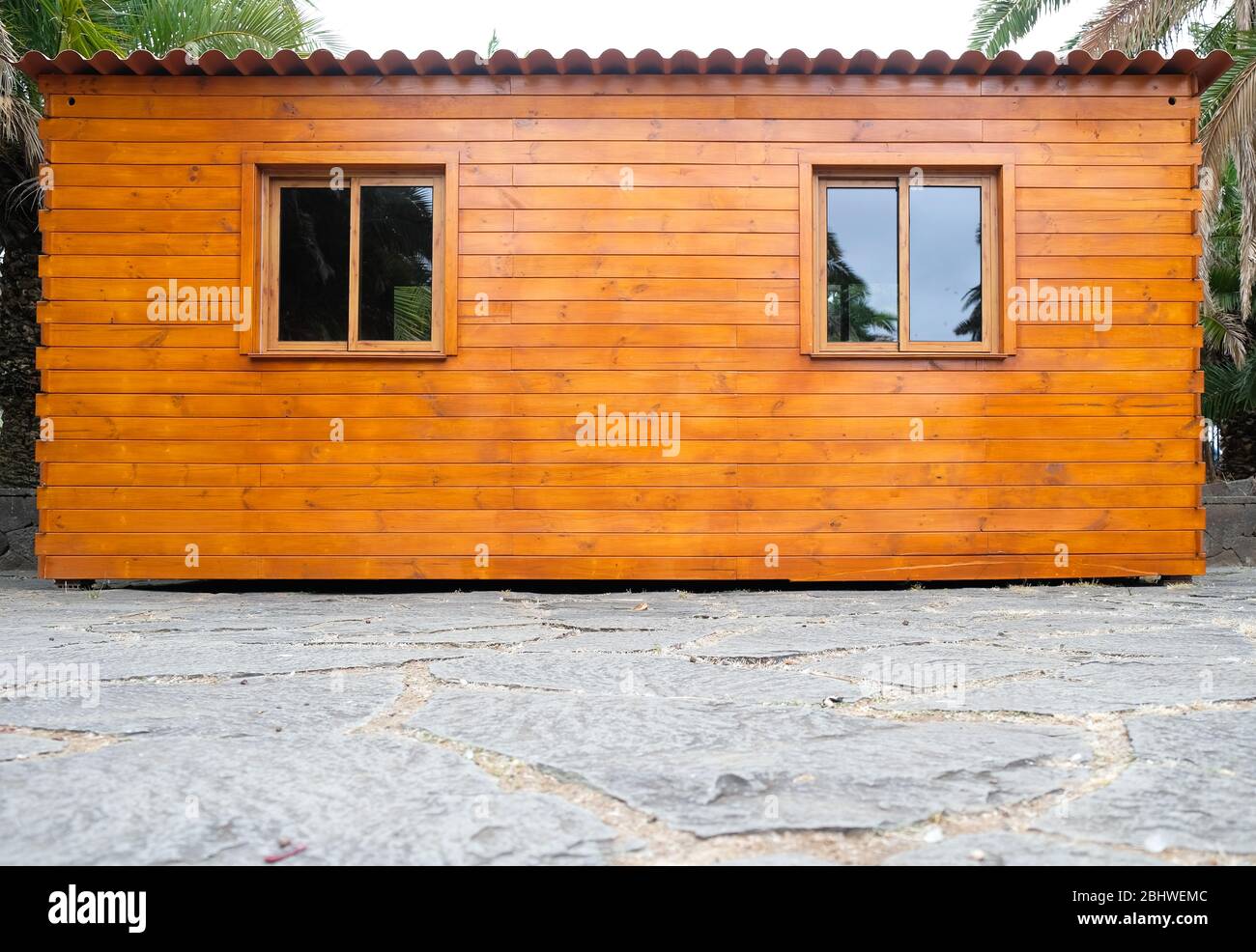 Cabina semplice in legno con finestre e tetto in ferro ondulato, circondata da palme, vista frontale. Foto Stock
