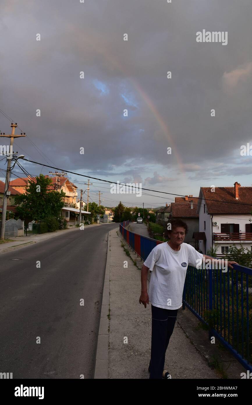 Sul marciapiede accanto alla strada, una donna più anziana si alza. Sullo sfondo sono le case e arcobaleni nel cielo Foto Stock