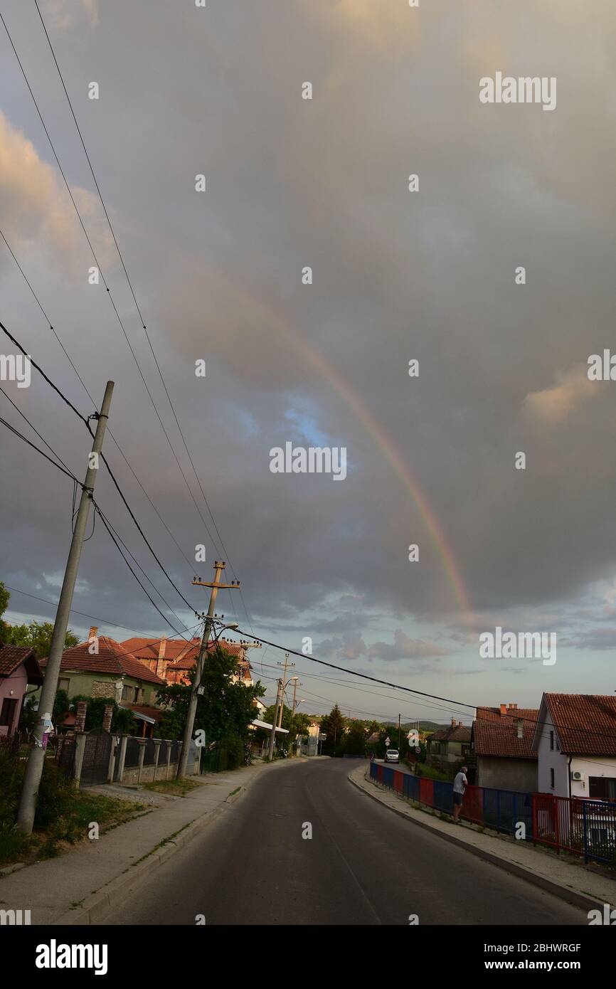 Nel cielo nuvole grigie con arcobaleno. Strada asfaltata passa attraverso il villaggio. Vicino alla strada ci sono case e poli elettrici Foto Stock