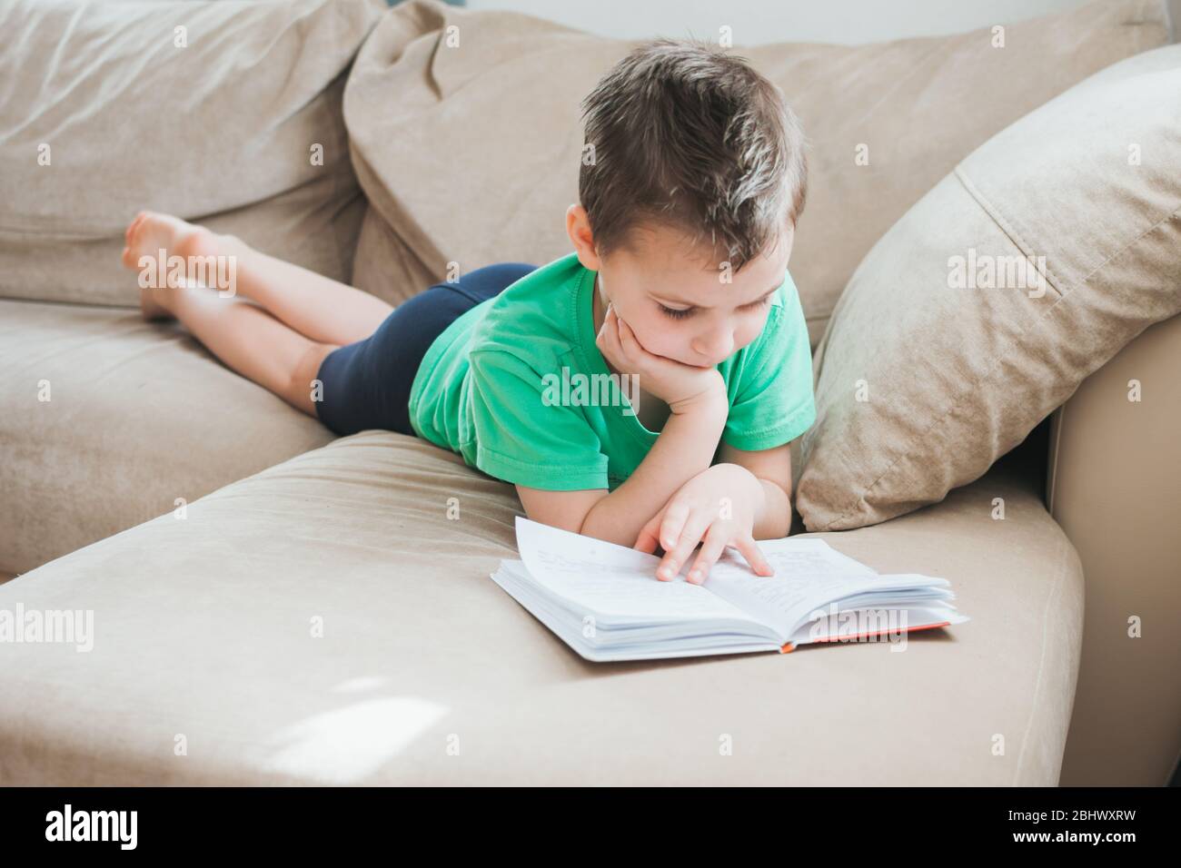 Ragazzo in maglietta verde leggendo un libro a casa sul divano. Formazione a distanza. Detox digitale. Ragazzo in quarantena rimanere a casa in isolamento auto. Foto Stock