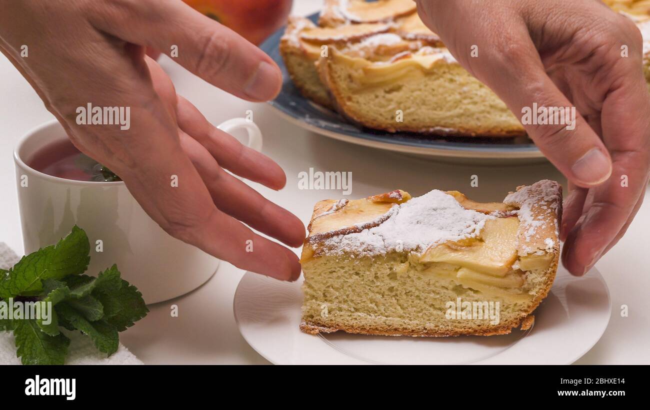 Fetta di torta di mele da vicino su un piatto, e tazza di tè alle erbe con foglie di menta da vicino sul tavolo da cucina bianco. Donna che serve torta. Foto Stock