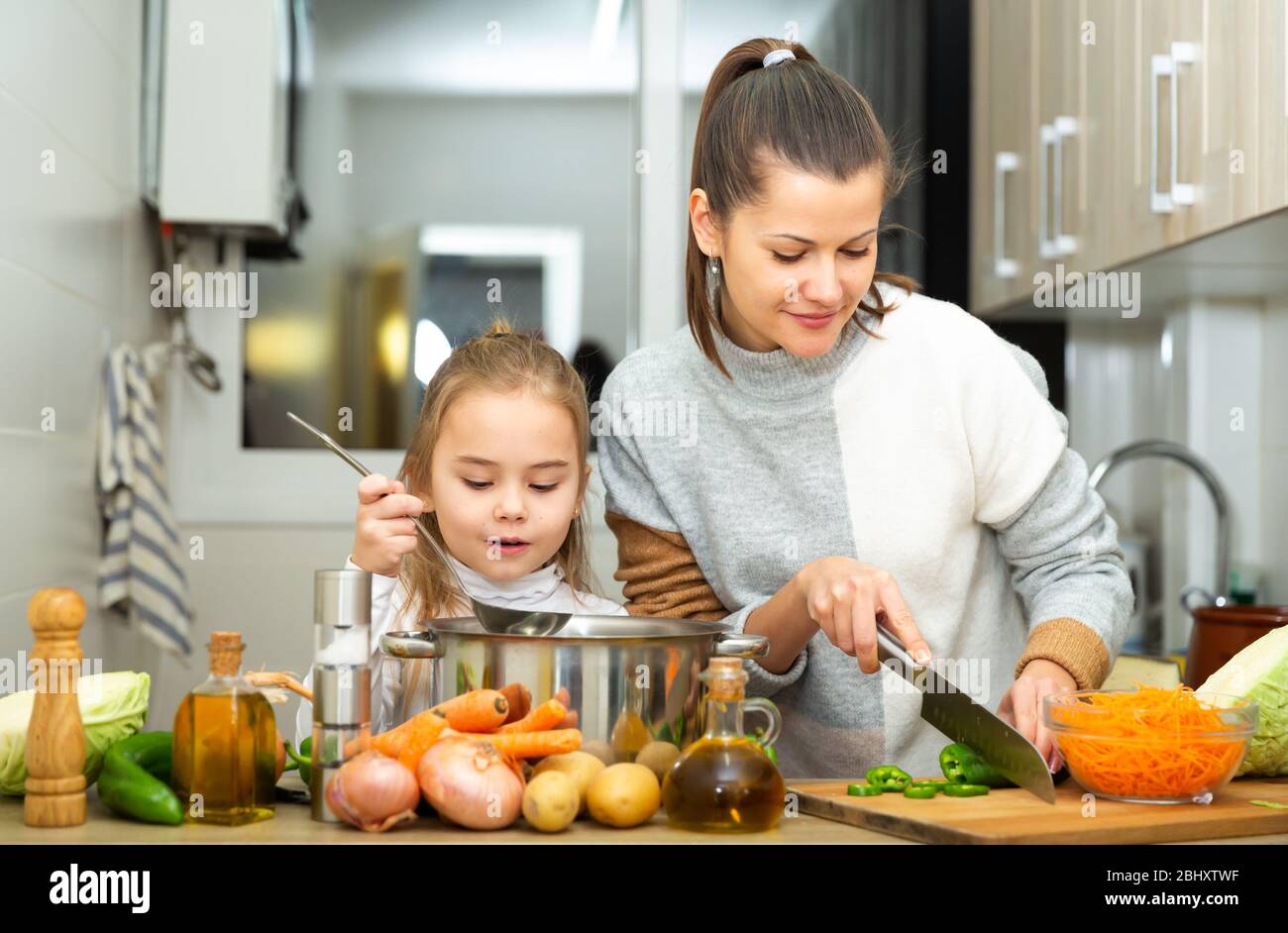 Madre felice e piccola figlia che preparano insieme la zuppa a casa, tagliano le verdure Foto Stock