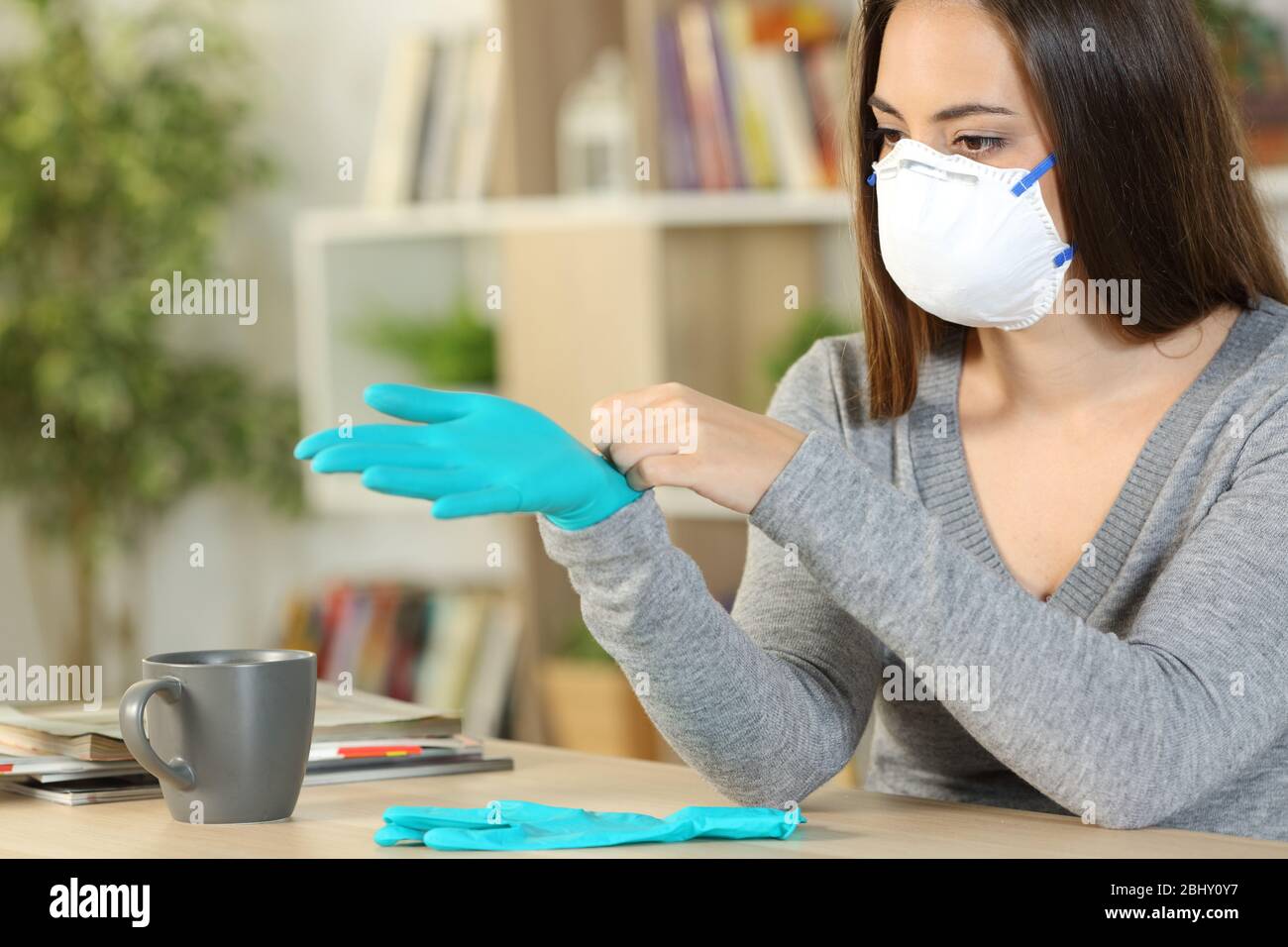 Donna con maschera protettiva mette i guanti di lattice evitando il contagio del coronavirus seduto su una scrivania a casa Foto Stock