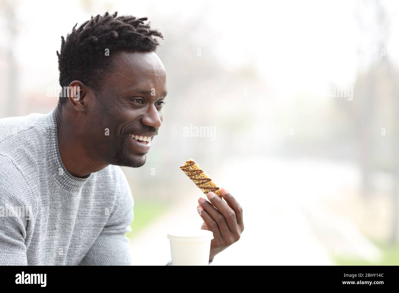 Un uomo nero felice che ha un caffè da asporto e uno snack bar di cereali in una panchina del parco Foto Stock
