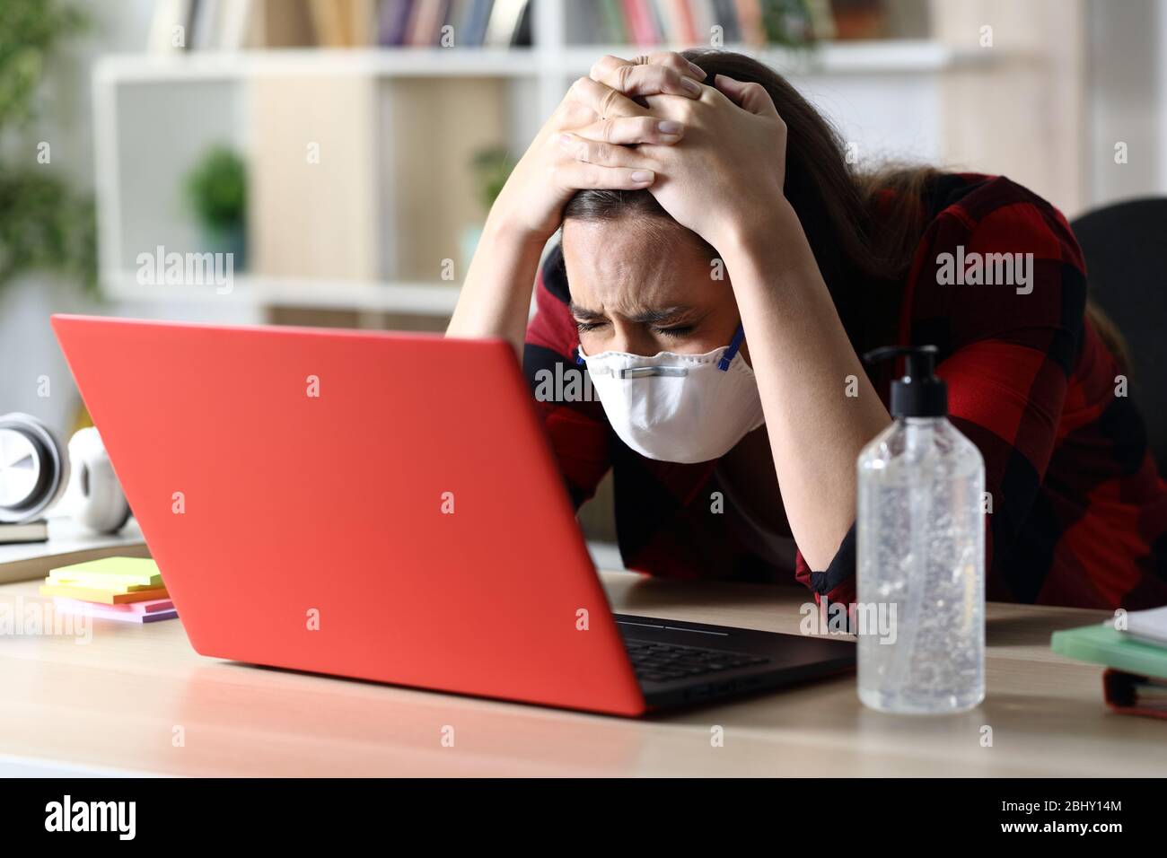 Donna studentesca triste con maschera protettiva in quarantena di coronavirus che controlla il laptop seduto su una scrivania a casa Foto Stock