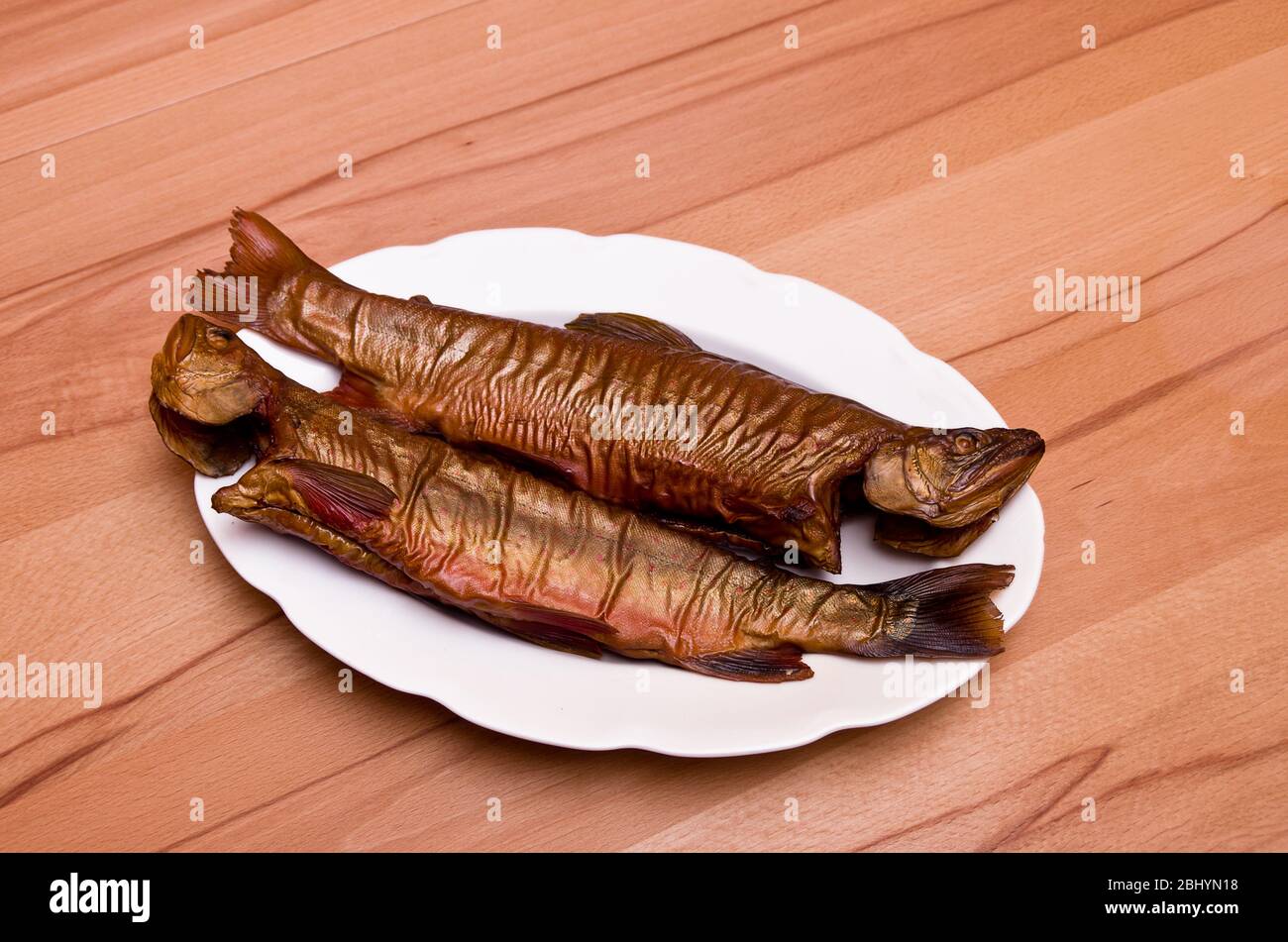 Due pesci affumicati, trota di ruscello, su un piatto bianco su un tavolo di faggio. Foto Stock
