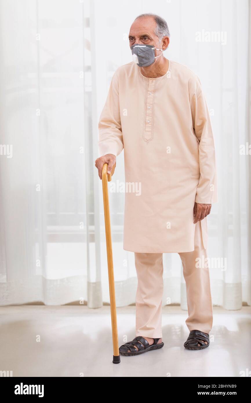 Uomo anziano che cammina con un bastone che indossa una maschera di inquinamento a casa. (Salute e fitness) Foto Stock