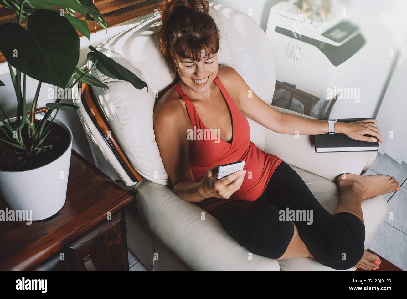 Affascinante donna di mezza età che guarda smartphone e sorridente, seduta con un libro seduto su una poltrona in un soggiorno. Foto Stock