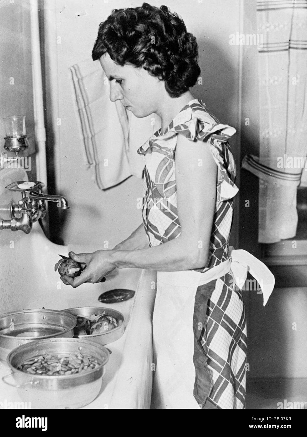 La signora William Grant Hayes, una casalinga cieca e sorda di Lymington, nel New Jersey, gestisce la sua casa in modo efficiente nonostante i suoi grandi svantaggi. Cucina i pasti per la sua famiglia di tre e può preparare facilmente il cibo per 20 ospiti. La onorevole Hayes si è formata a condurre una conversazione con le mani. La persona con cui parla usa il dito come penna e scrive sul palmo. La signora William Grant Hayes che spellano le patate nella sua casa a Lymington, New Jersey. - 27 agosto 1937 Foto Stock