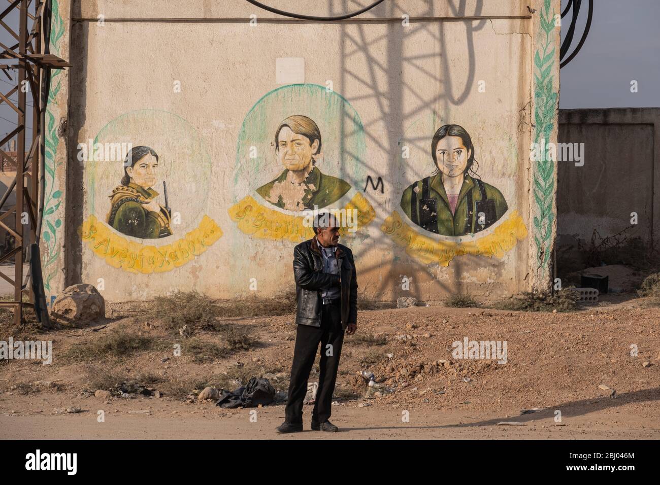 Un uomo attende dal lato della strada con un murale che indica il sacrificio di donne combattenti curdi durante il conflitto siriano dietro di lui. Qamishli, Siria. Foto Stock