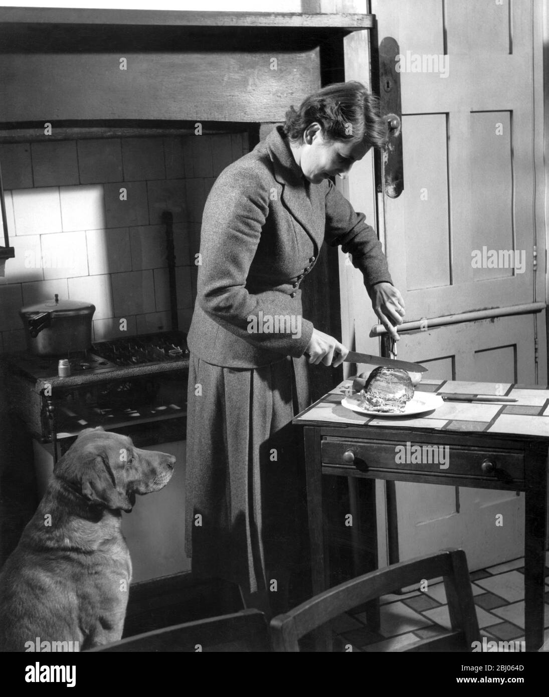 Casalinga intagliare la carne sulla tavola da cucina come il cane di famiglia guarda con interesse. 1952 Foto Stock