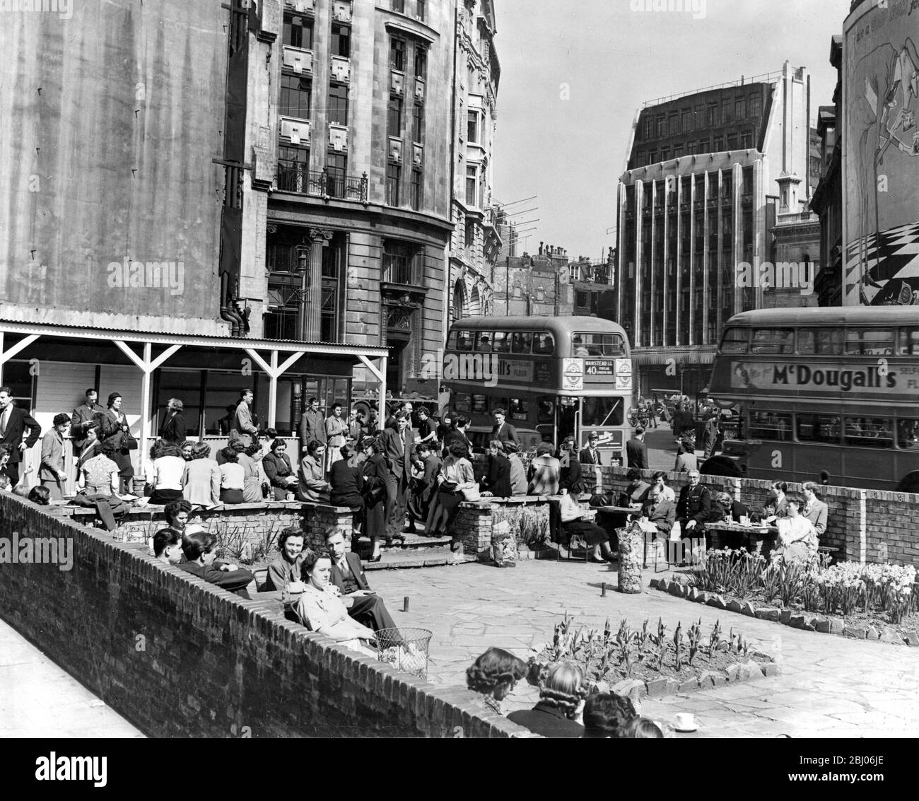 Il caldo sole luminoso è venuto a Londra e coloro che possono approfittarne sono lieti di questa opportunità. Per coloro che sono confinati agli uffici tutto il giorno è frustrante, ma un po' di aiuto è il piacevole giardino da tè attaccato ad un caffè in GT William Street, vicino al Monument, dove i lavoratori della città possono rilassarsi e gustare i loro spuntini al sole. La foto mostra una vista del piacevole giardino del tè in GT William Street, con gli impiegati che si rilassano mentre il traffico di Londra è molto trafficato. 18 aprile 1952 Foto Stock