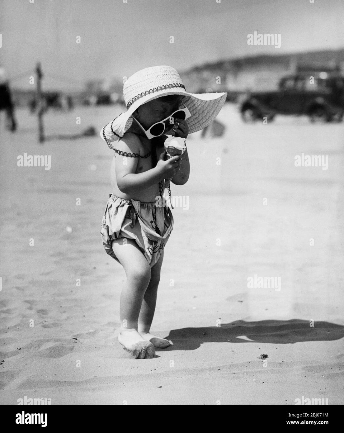 Piccolo bambino che cerca di mangiare un cono gelato sulla sabbia calda. Agosto 1949 - Foto Stock