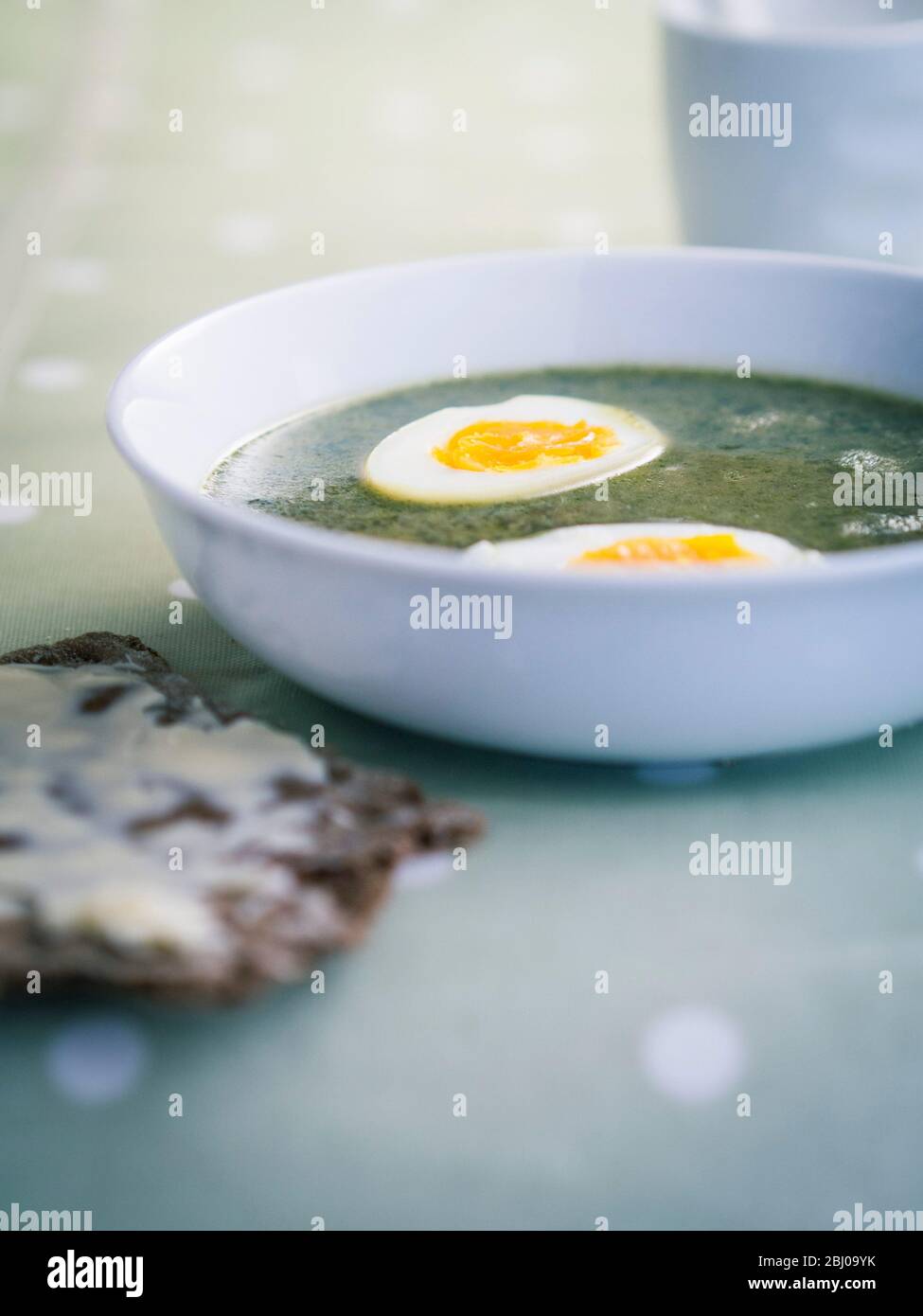 Ciotola bianca di zuppa di ortica svedese su panno a righe con cucchiaio d'argento su panno di verdure macchiato con knÌÄå ckebrÌÄå¦d svedese (pane croccante di segale e bu Foto Stock