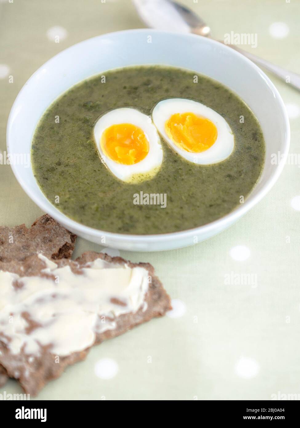 Ciotola bianca di zuppa di ortica svedese su panno a righe con cucchiaio d'argento su panno di verdure macchiato con knÌÄå ckebrÌÄå¦d svedese (pane croccante di segale e bu Foto Stock