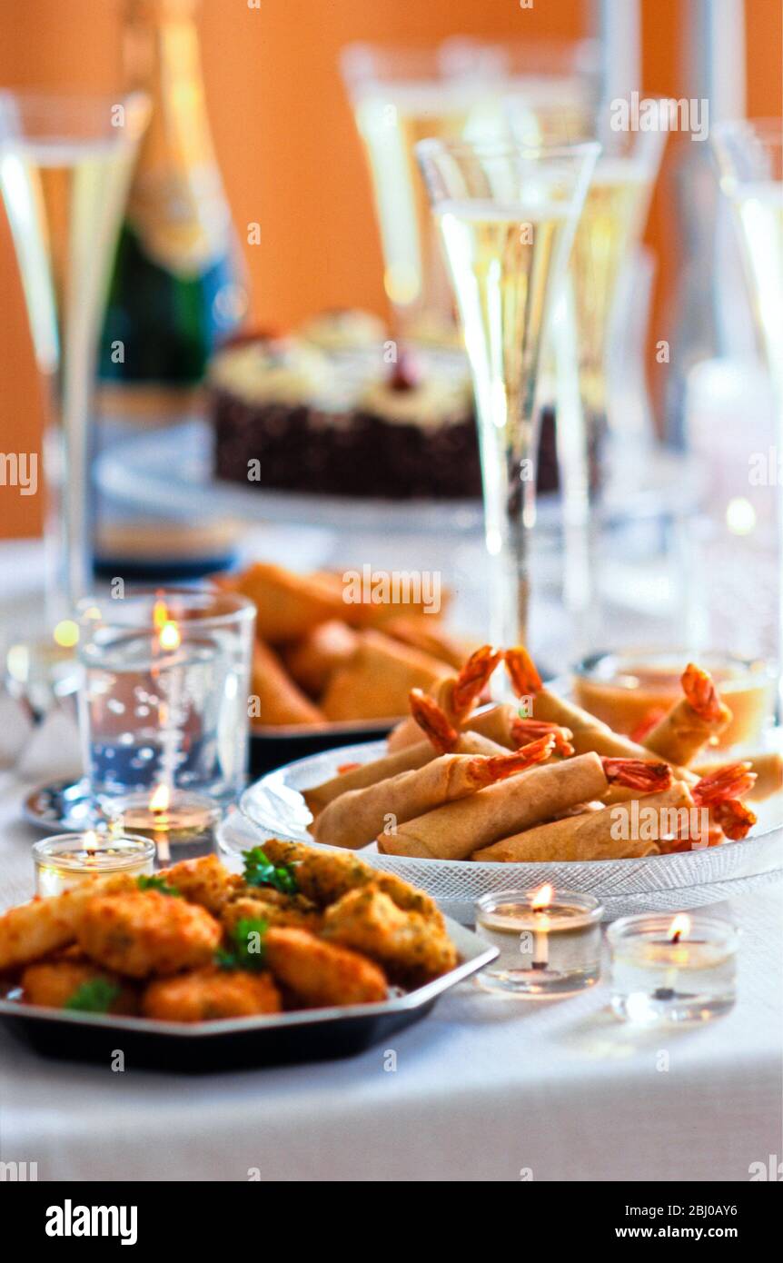 Tavolo con tartine di festa (involtini di primavera di gamberi re e mini torte di pesce thailandesi) e alti flauti di champagne con torta di Natale in background - Foto Stock