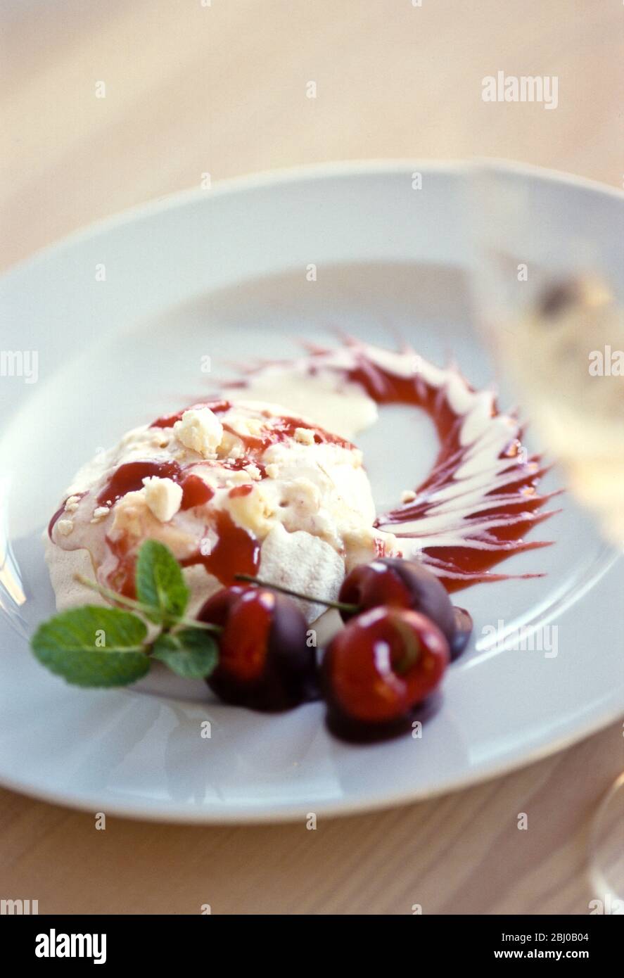 Spettacolare, dessert di meringa con gelato alla vaniglia, coulis di ciliegia e ciliegie fresche immerse in cioccolato fondente su piatto bianco guarnito con menta Foto Stock