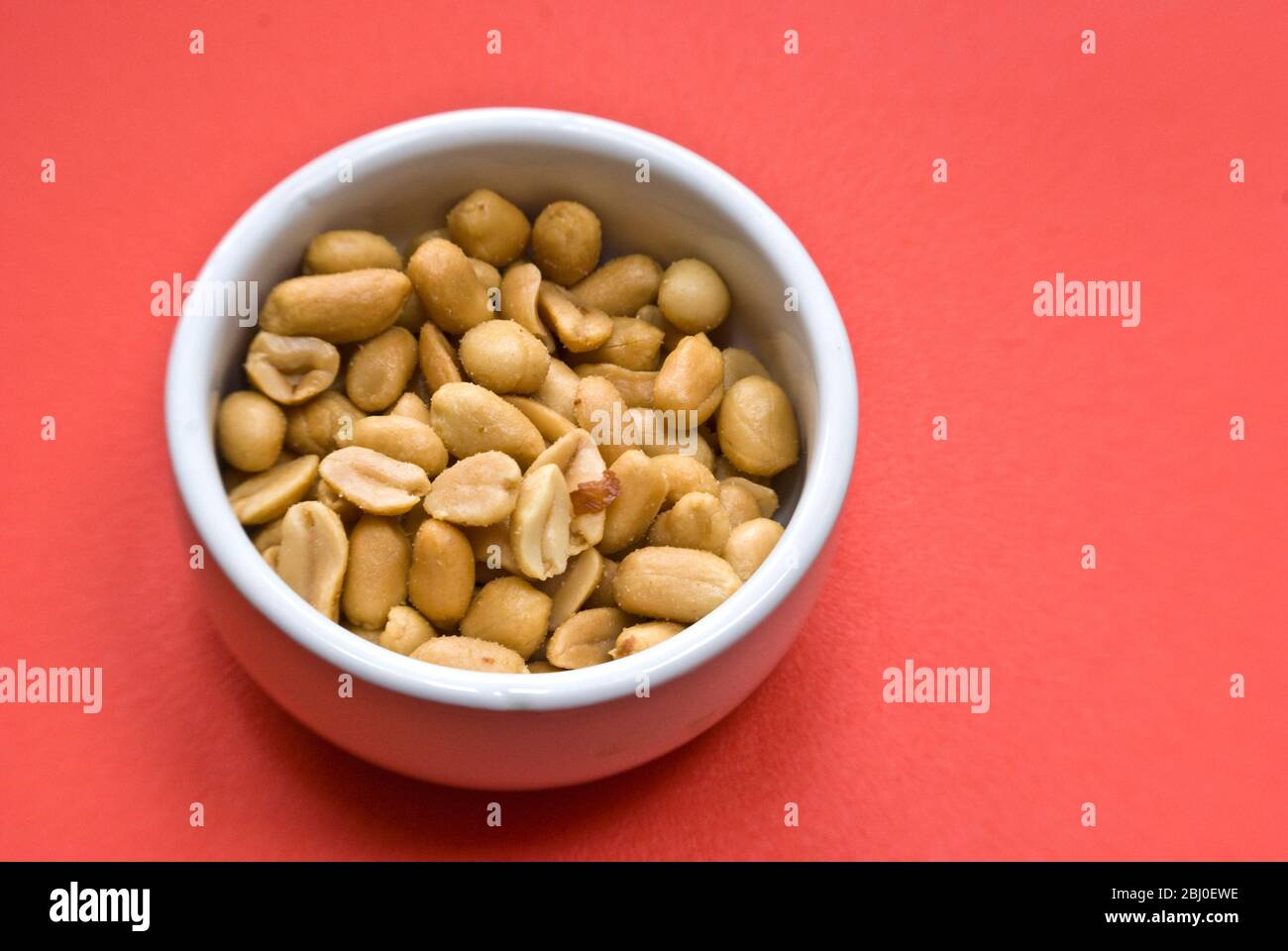 Piccola ciotola bianca di arachidi salate e arrostite su fondo rosso brillante - Foto Stock