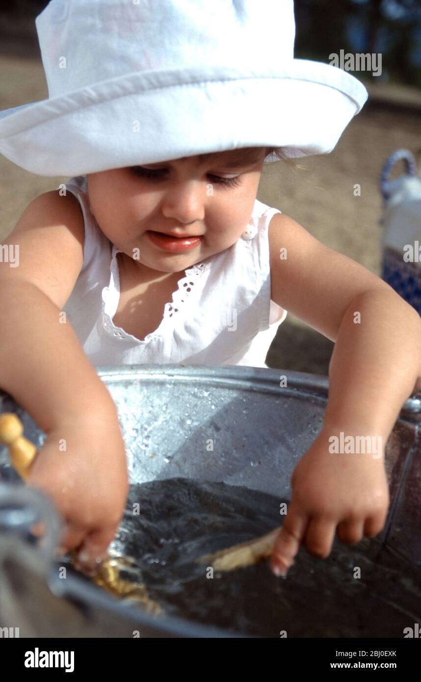 Bambina che gioca con un secchio di acqua limpida e alcuni pioli di vestiti mentre la madre sta appendendo fuori il lavaggio in cortile soleggiato - Foto Stock