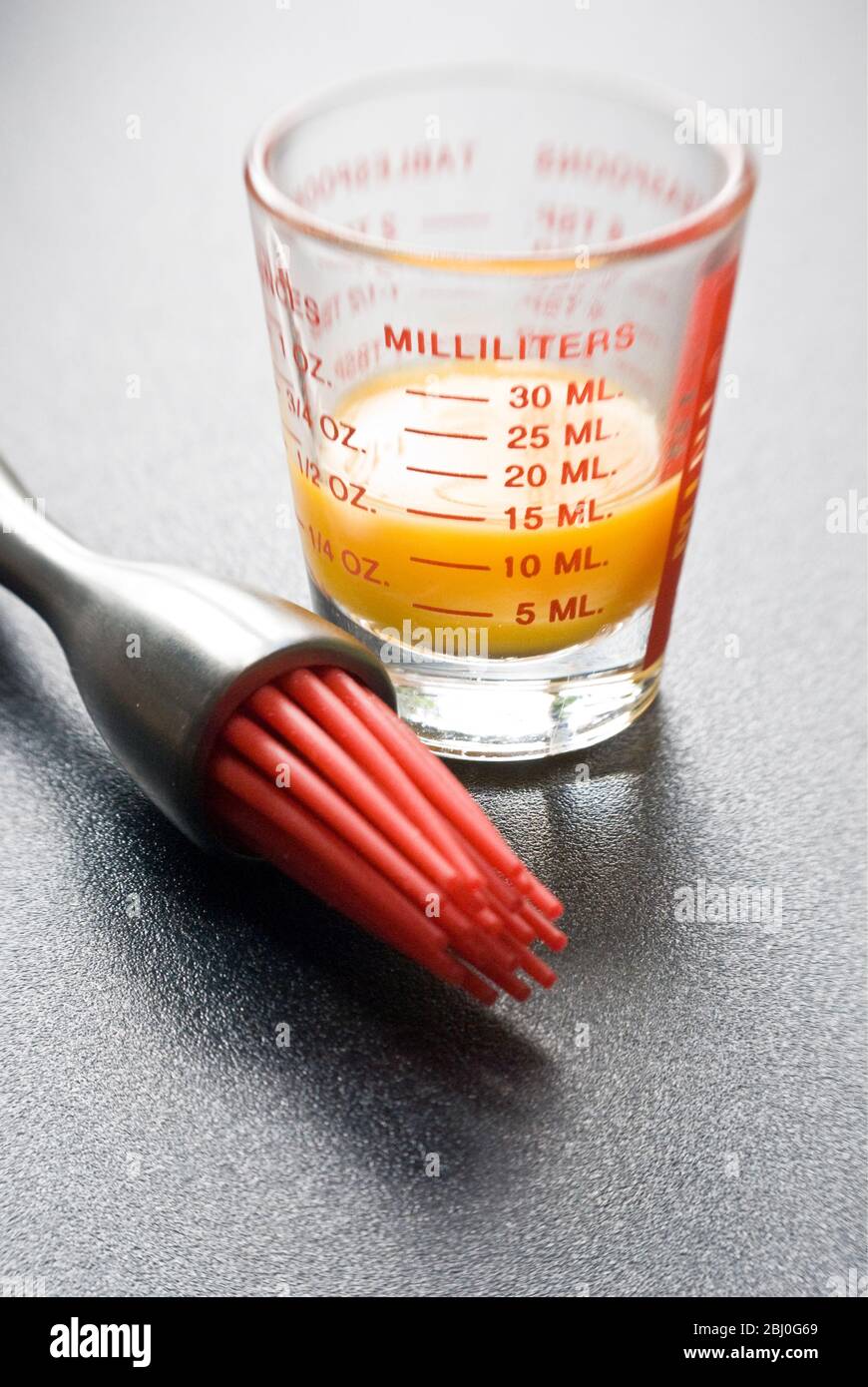 Tuorlo d'uovo in piccolo bicchiere graduato con moderno pennello da pasticceria con 'setole' in silicone rosso - Foto Stock