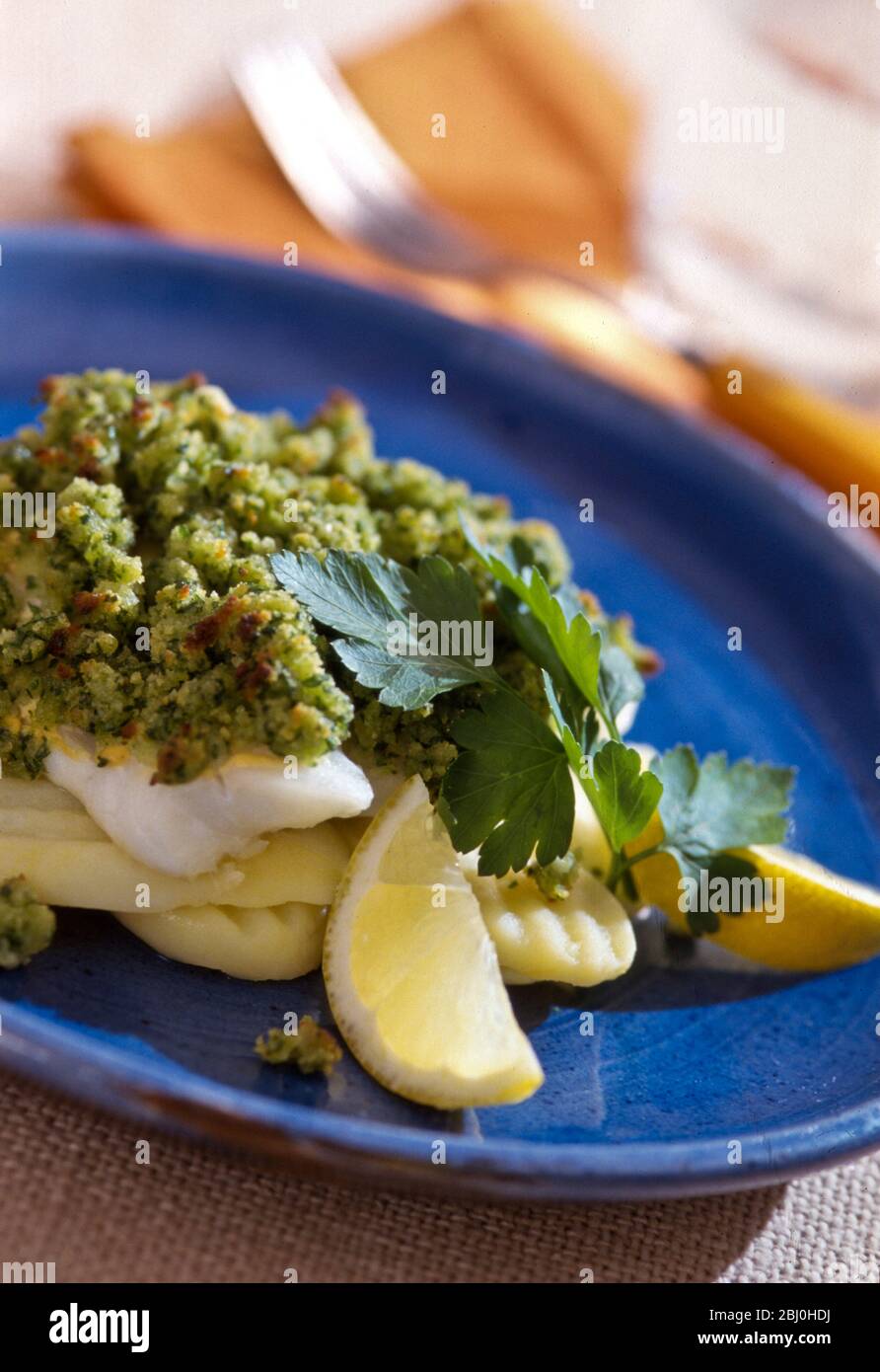 Pesce con crosta di prezzemolo su patate tagliate al vapore, servite con spicchi di limone su piastra blu. - Foto Stock