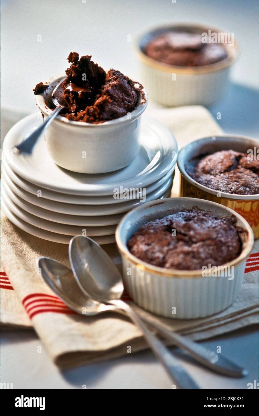 Budini di cioccolato caldo in piccoli ramekins, con il ppile di piatti, panno rosso e bianco e cucchiai - Foto Stock