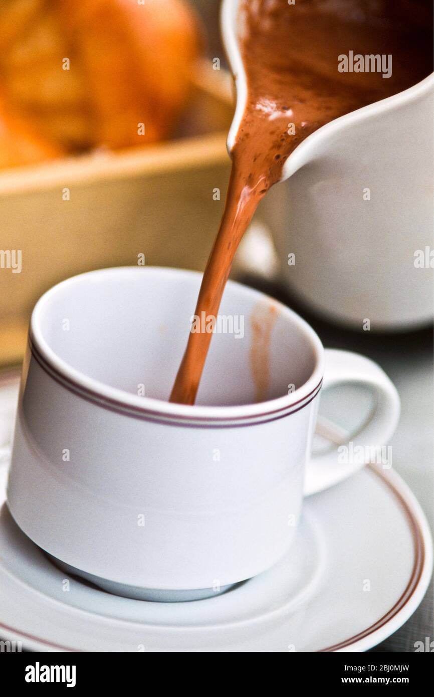 Versare il cioccolato caldo dalla brocca in tazza e piattino sul tavolo del caffè, Francia, con brioche nel cestino dietro - Foto Stock