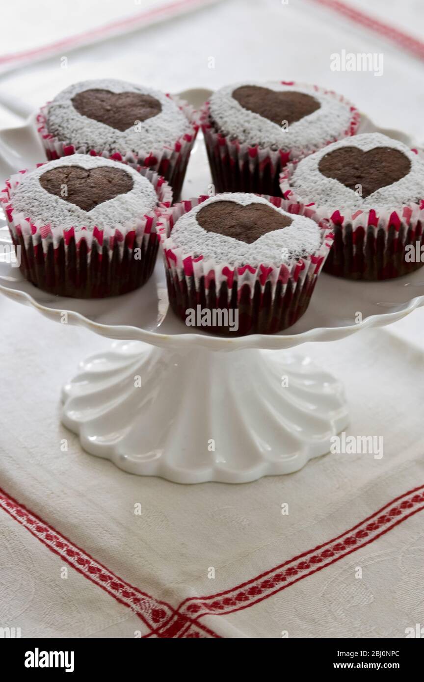 Cinque muffin al cioccolato decorati con zucchero a velo a forma di cuore su base per torta bianca - Foto Stock