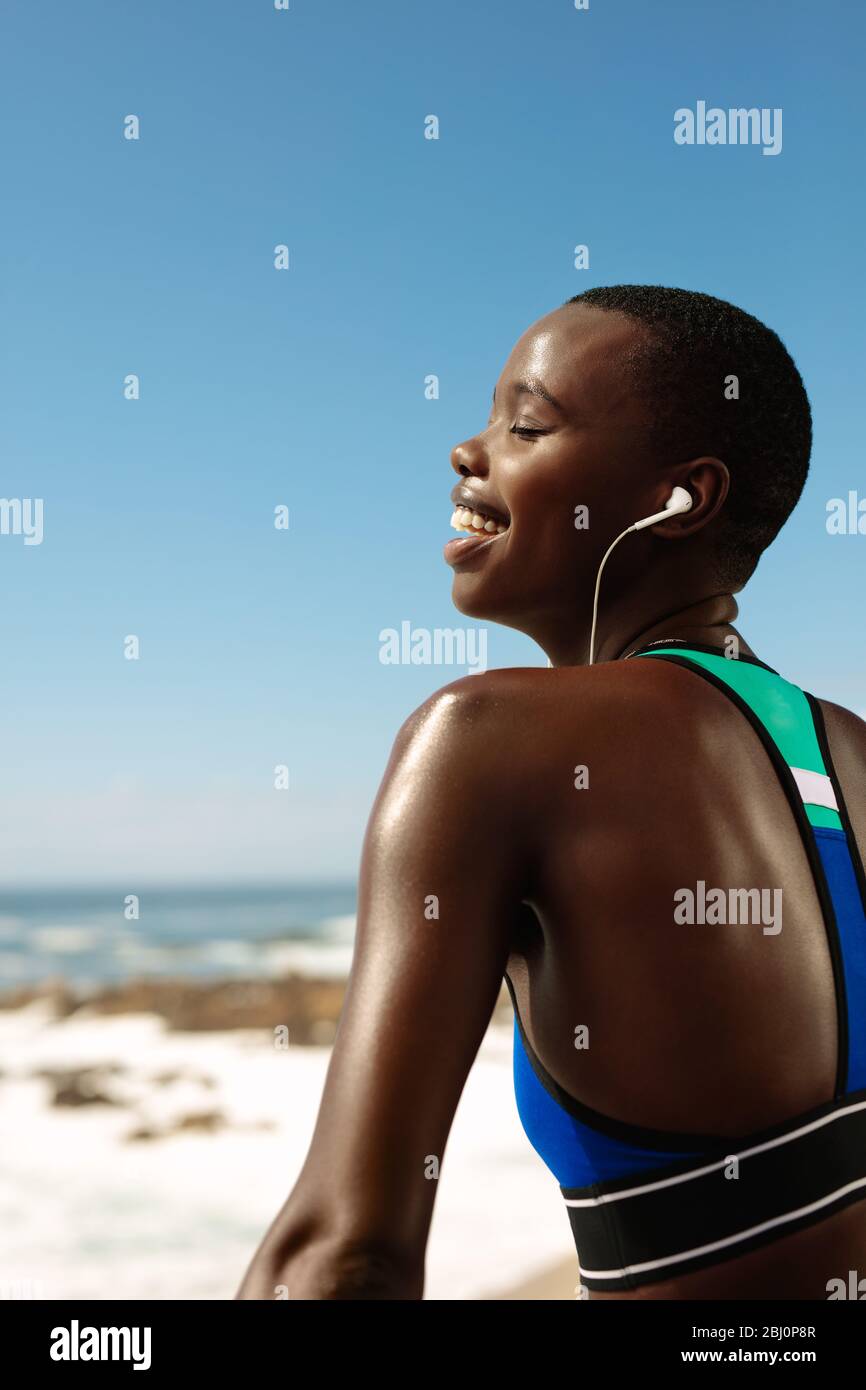 Donna fitness che indossa gli auricolari ascoltando musica e sorridendo. Donna africana in abbigliamento sportivo prendendo una pausa dopo l'esercizio all'aperto. Foto Stock