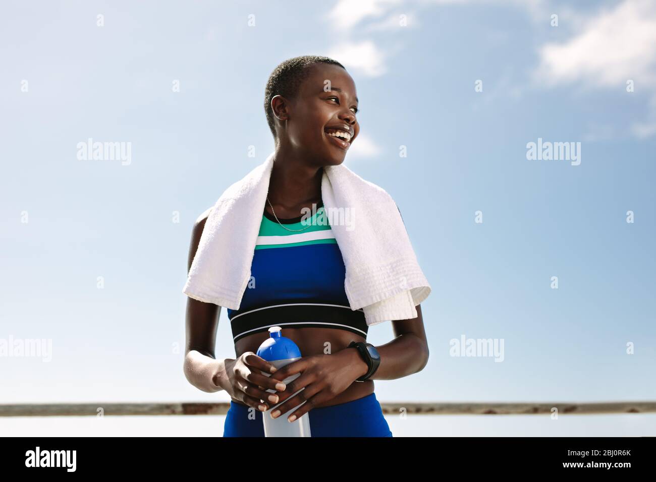 Donna sportiva con asciugamano intorno al collo e tenendo una bottiglia d'acqua che guarda via e sorridente. Atleta femminile che prende pausa dopo sessione di allenamento all'aperto Foto Stock