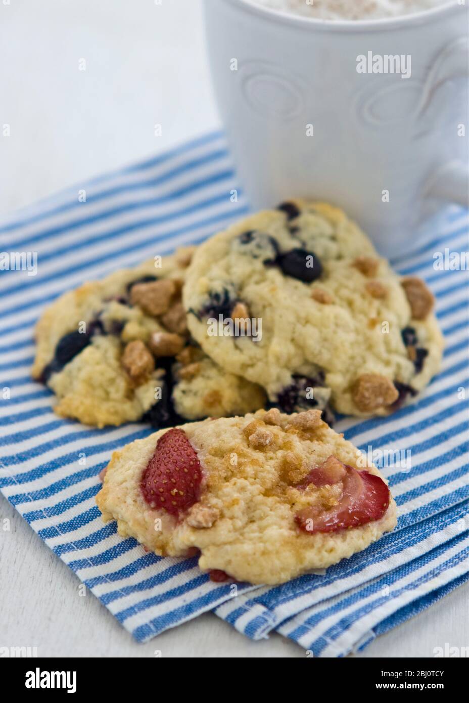 Biscotti morbidi con frutti di bosco e noci su tovagliolo a righe bianche e blu con tazza di cappuccino - Foto Stock