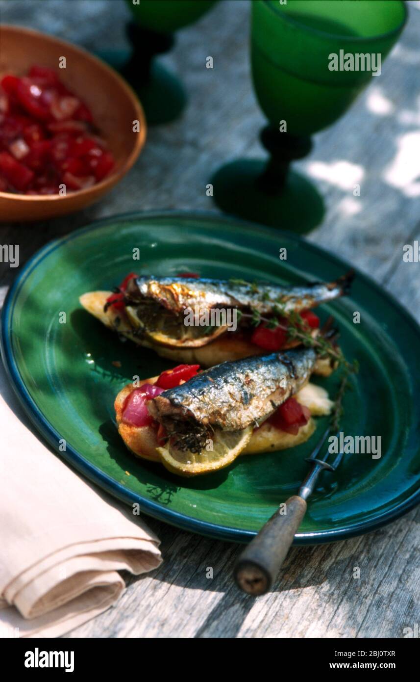 Fresche grigliate, sardine con cipolla rossa e salsa di pomodoro su pane tostato su piatto verde su vecchio tavolo logoro fuori al sole - Foto Stock