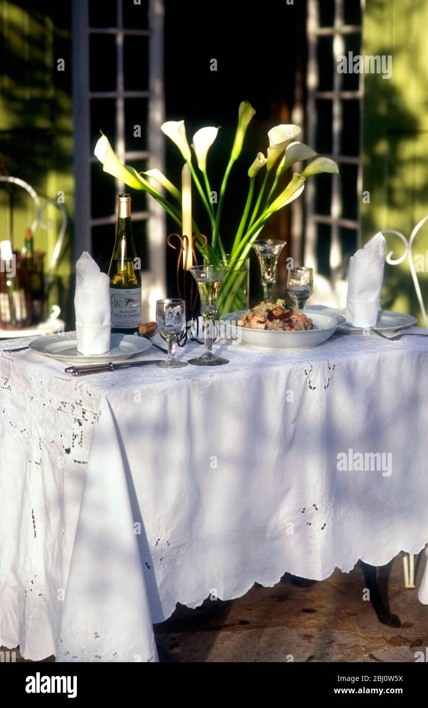 Bella tavola posata all'esterno con tela di merletto e gigli di arum bianco in luce del sole della sera fuori casa francese con persiane verdi - Foto Stock