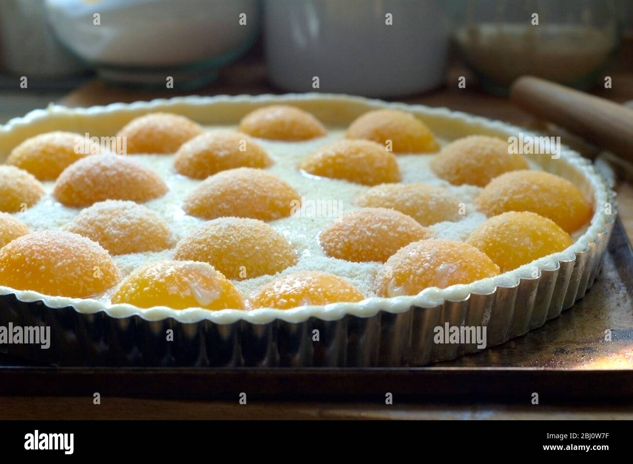 Preparazione della crostata di pesca con pesche in scatola dimezzate e crema patissiere su pasta sfoglia in latta di metallo, cosparso di zucchero di caster e mandorle macinate. - Foto Stock