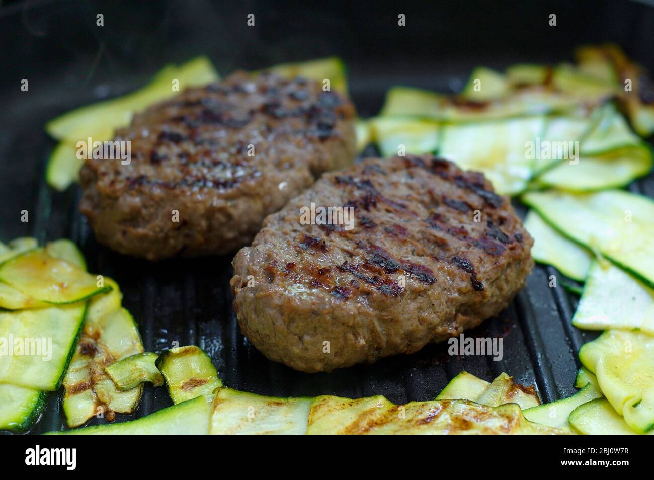 Hamburger di cervo grigliati su griddle caldo con scivoli di zucchine condite con olio d'oliva che viene cotto intorno a loro - Foto Stock