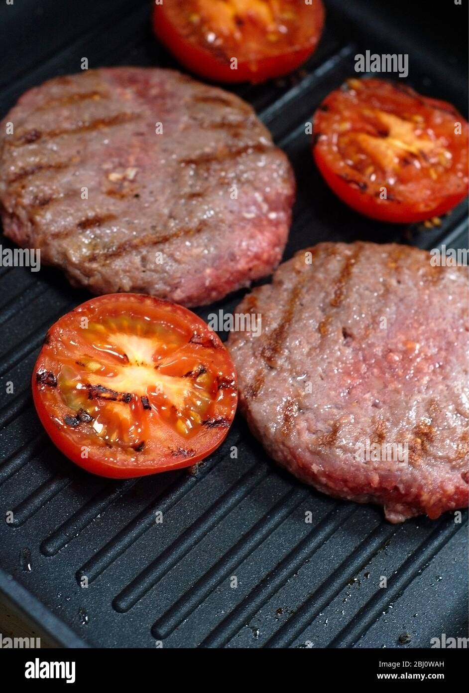 Hamburger e pomodori dimezzati alla griglia su una padella antiaderente - metà cotti, ancora rosa - Foto Stock