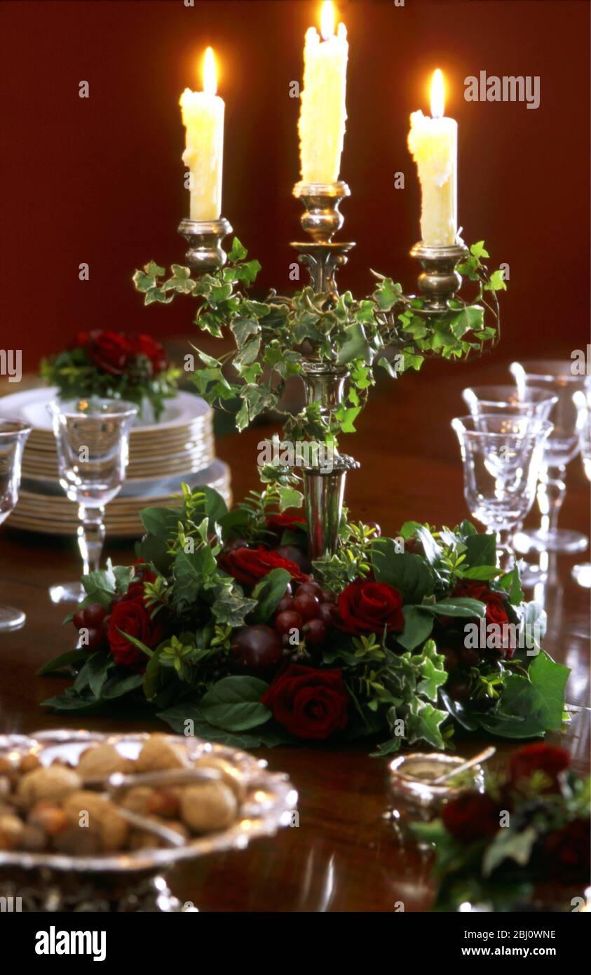 Formale rable pranzo di natale con candelabro decorato edera, vassoio di argento di noci, vetri di cristallo, sale, pila di piatti, e disposizione rosa rossa - Foto Stock