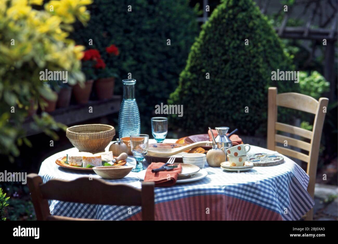 Tavolo pranzo all'aperto in giardino al sole, con sedie francesi e tovaglia a scacchi - Foto Stock
