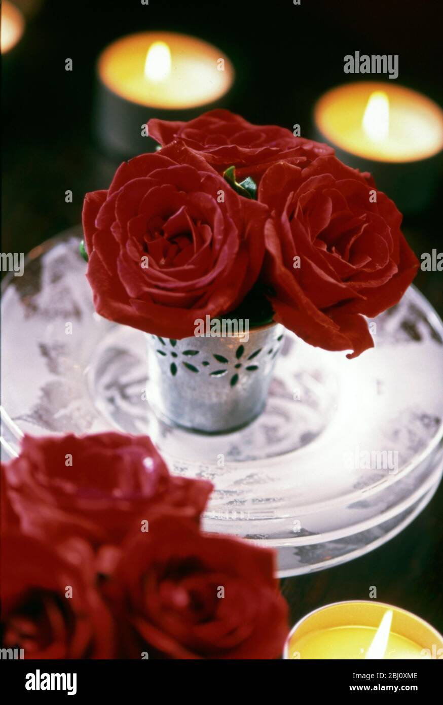 Tre rose rosse in piccolo vaso decorativo di metallo su pila di tre piatti rosati di vetro con candela tealights intorno. Decorazione tavola di Natale - Foto Stock