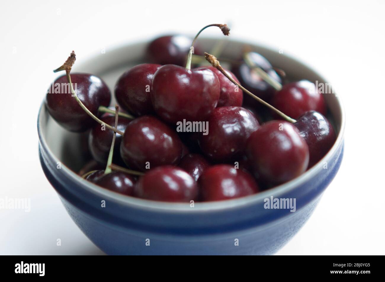 Una piccola ciotola blu di ciliegie rosse dolci su superficie bianca - Foto Stock