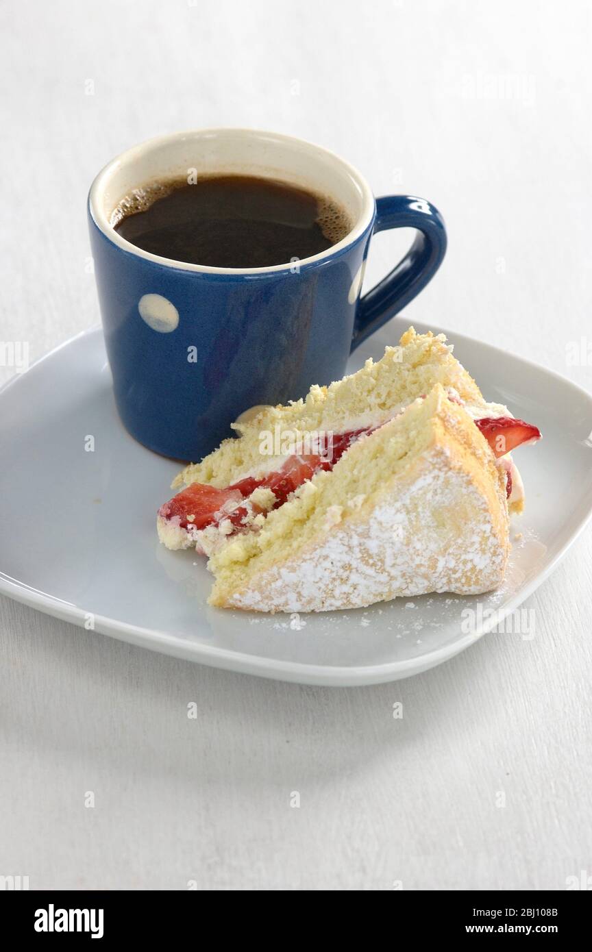 Un pezzo di torta di Spagna di fragola su un piatto bianco con una piccola tazza macchiata blu e bianca di caffè nero - Foto Stock