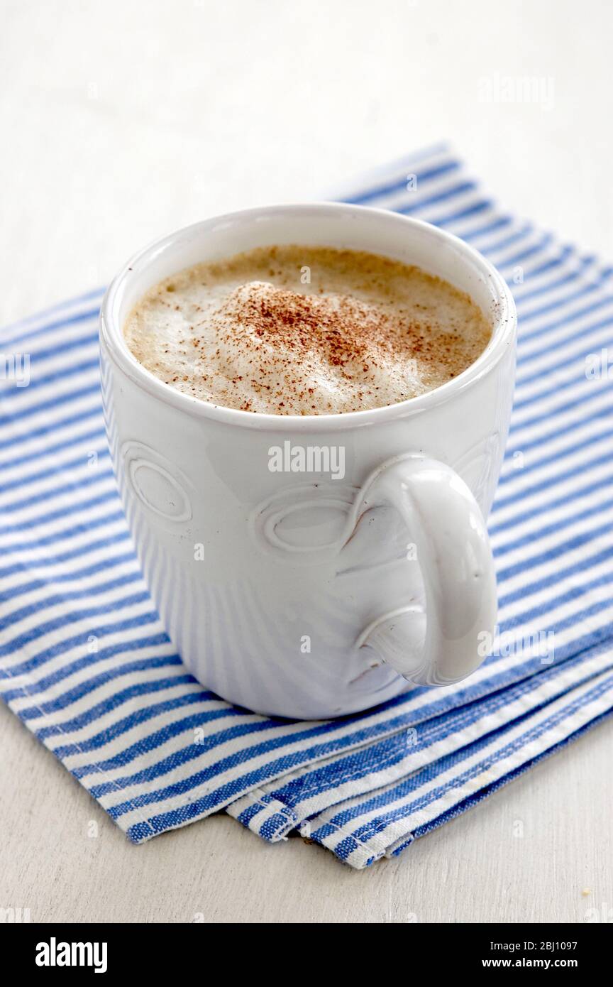 Tazza bianca di caffè cappuccino con farcitura di schiuma e polvere di cacao spruzzata su tovagliolo a righe blu e bianco - Foto Stock