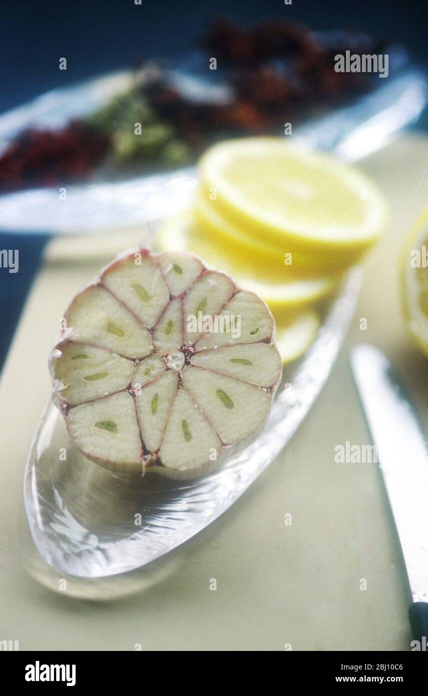 Bulbo d'aglio tagliato trasversalmente con fette di limone in un piatto di vetro su tagliere preparato per cucinare - Foto Stock